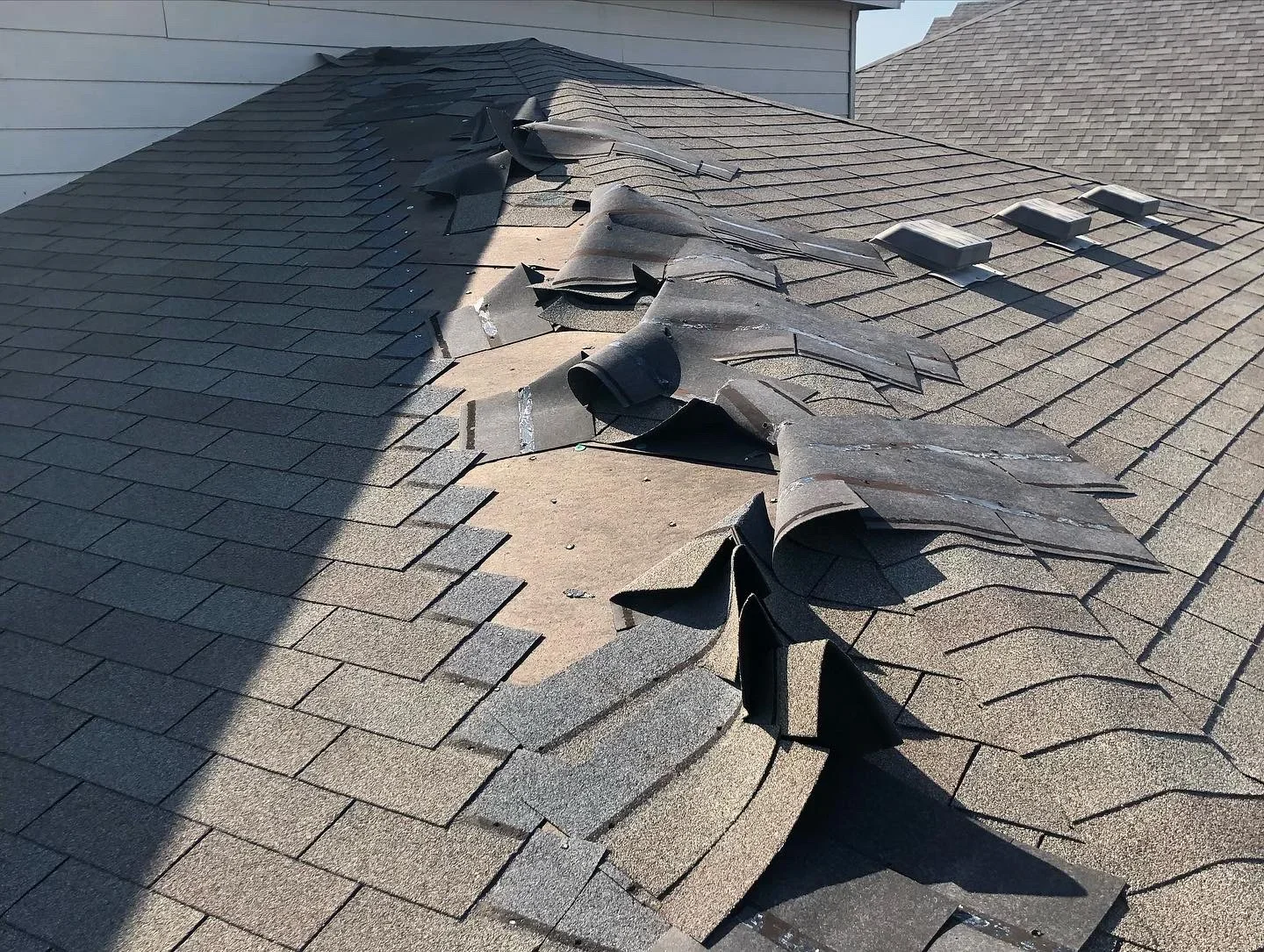 Damaged asphalt shingles with lifted and torn sections exposing roof decking during storm damage repair