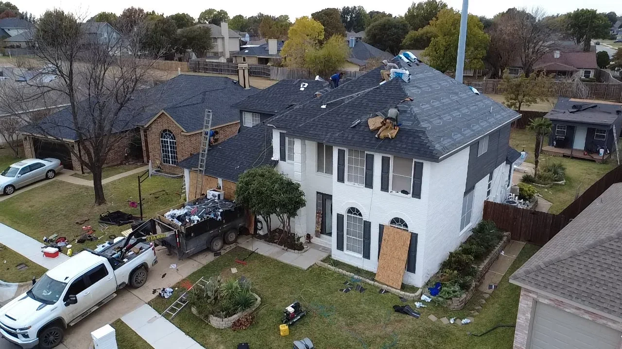 Two-story home with new charcoal gray architectural shingles being installed, workers on roof, equipment staged below