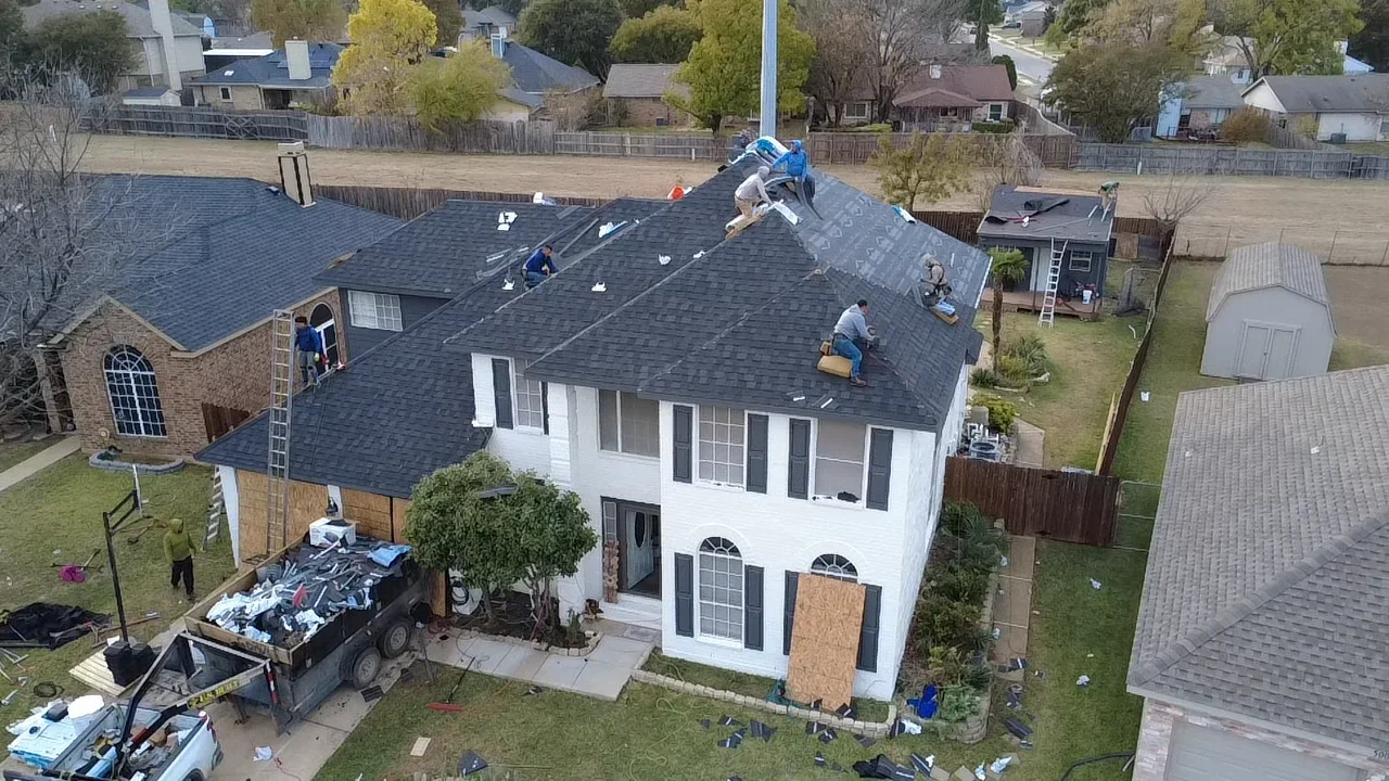 Roofers installing charcoal gray asphalt shingles on two-story home roof with crew and equipment visible