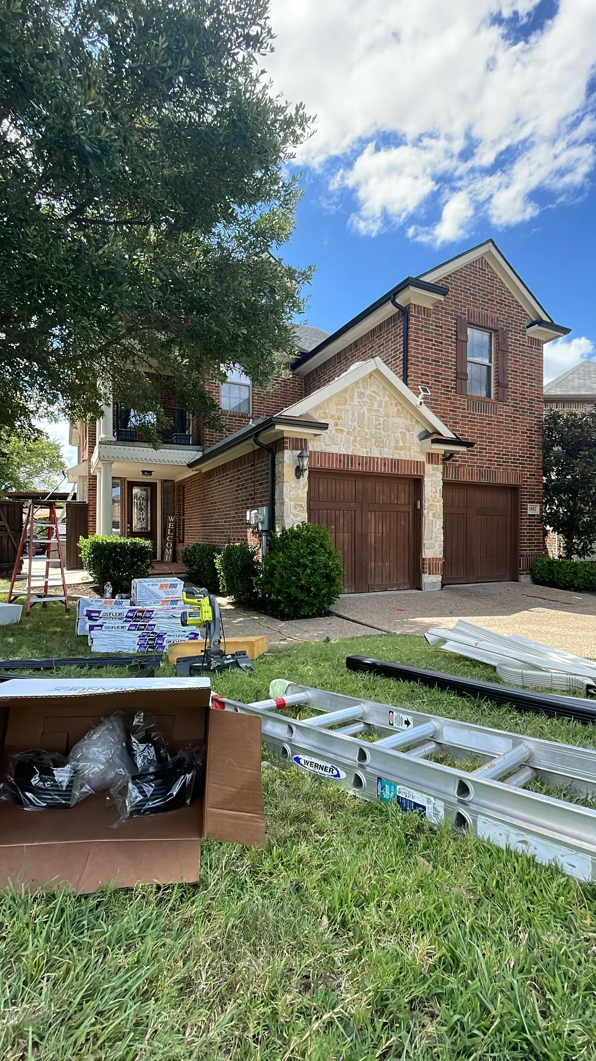Two-story brick home with new roof installation in progress, showing ladders and materials on lawn