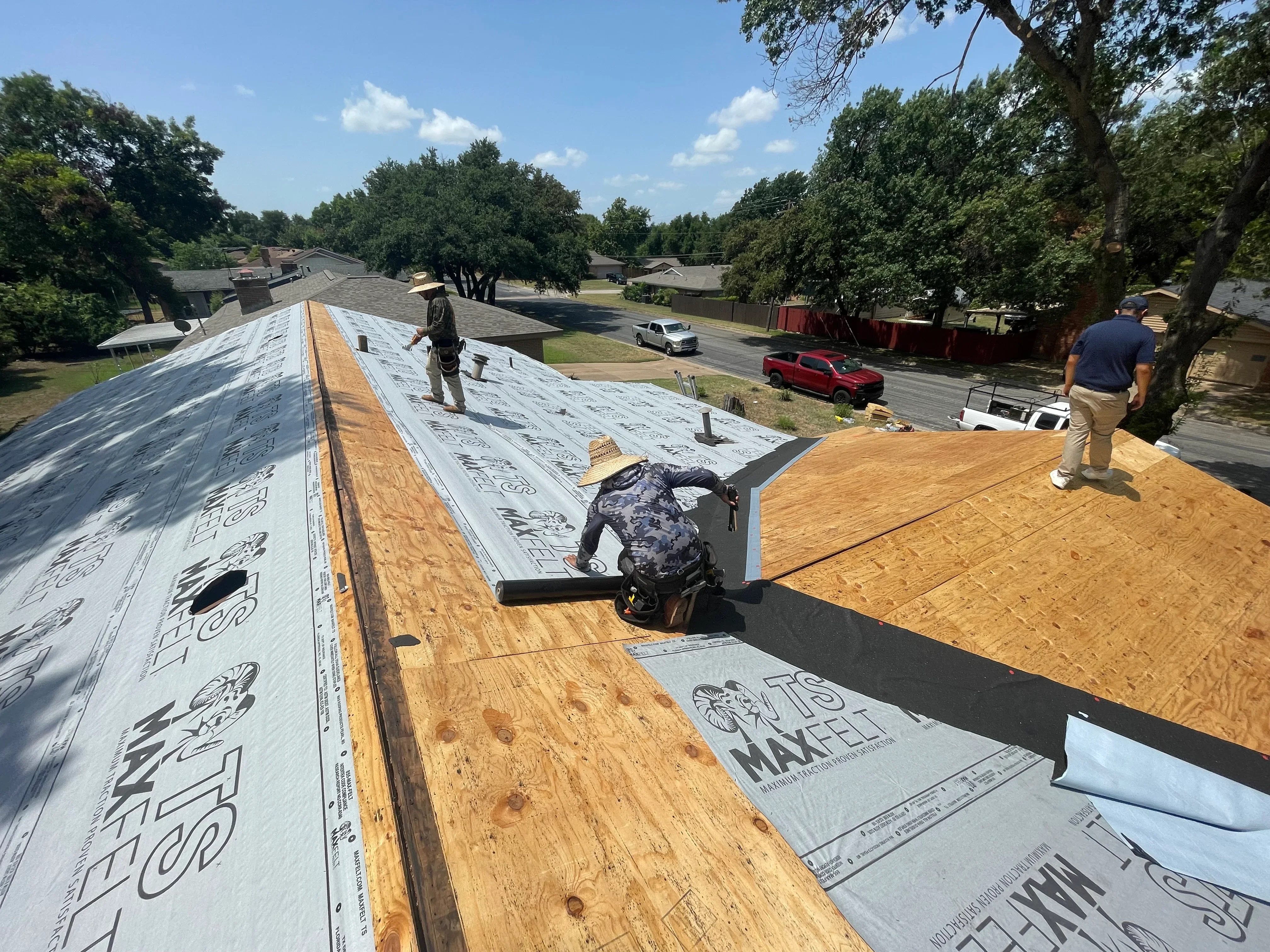 Roof replacement in progress with underlayment, plywood decking, and workers installing roofing materials